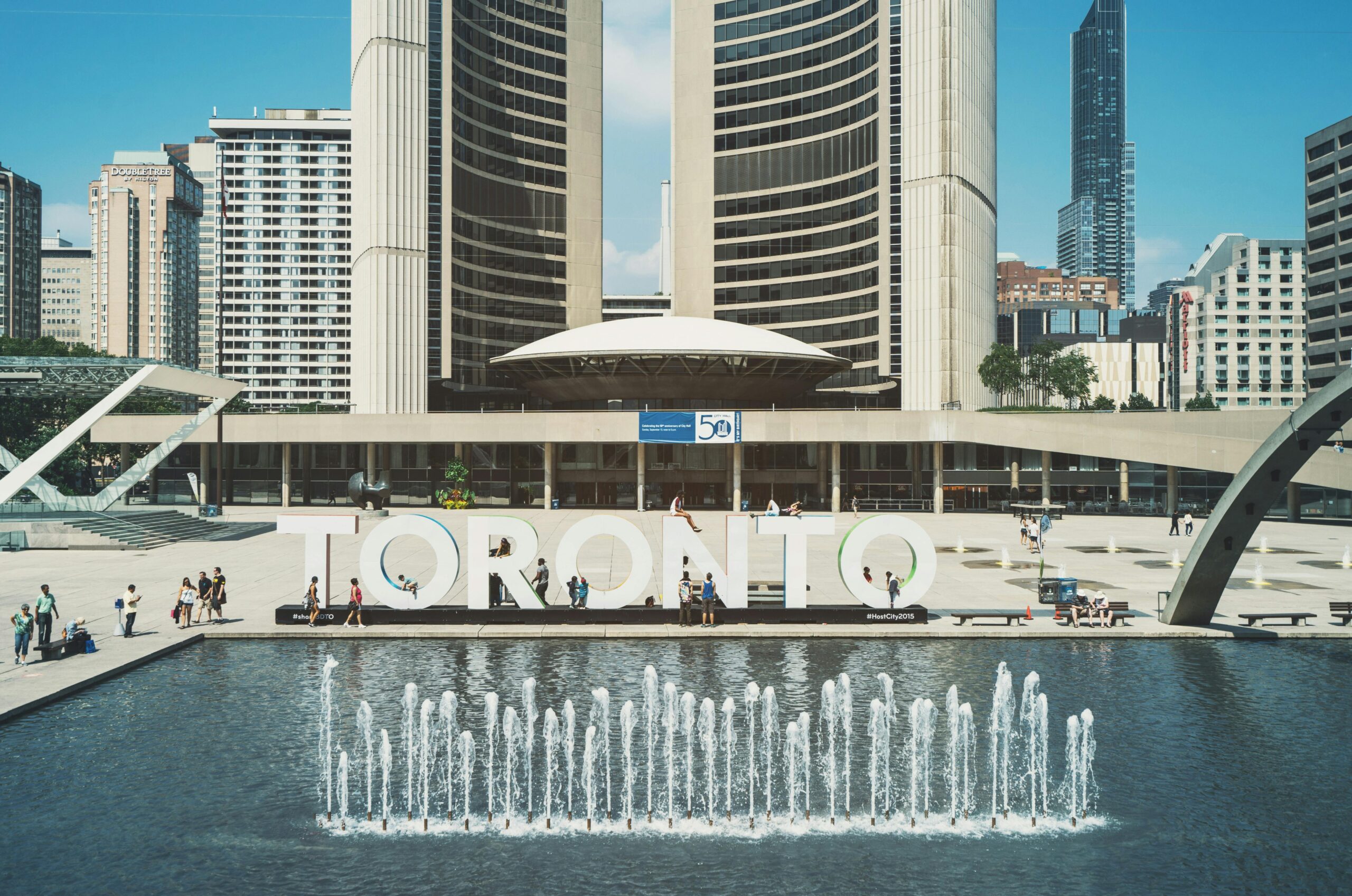 View of Toronto City Hall with vibrant fountains and iconic city sign under a blue sky.
