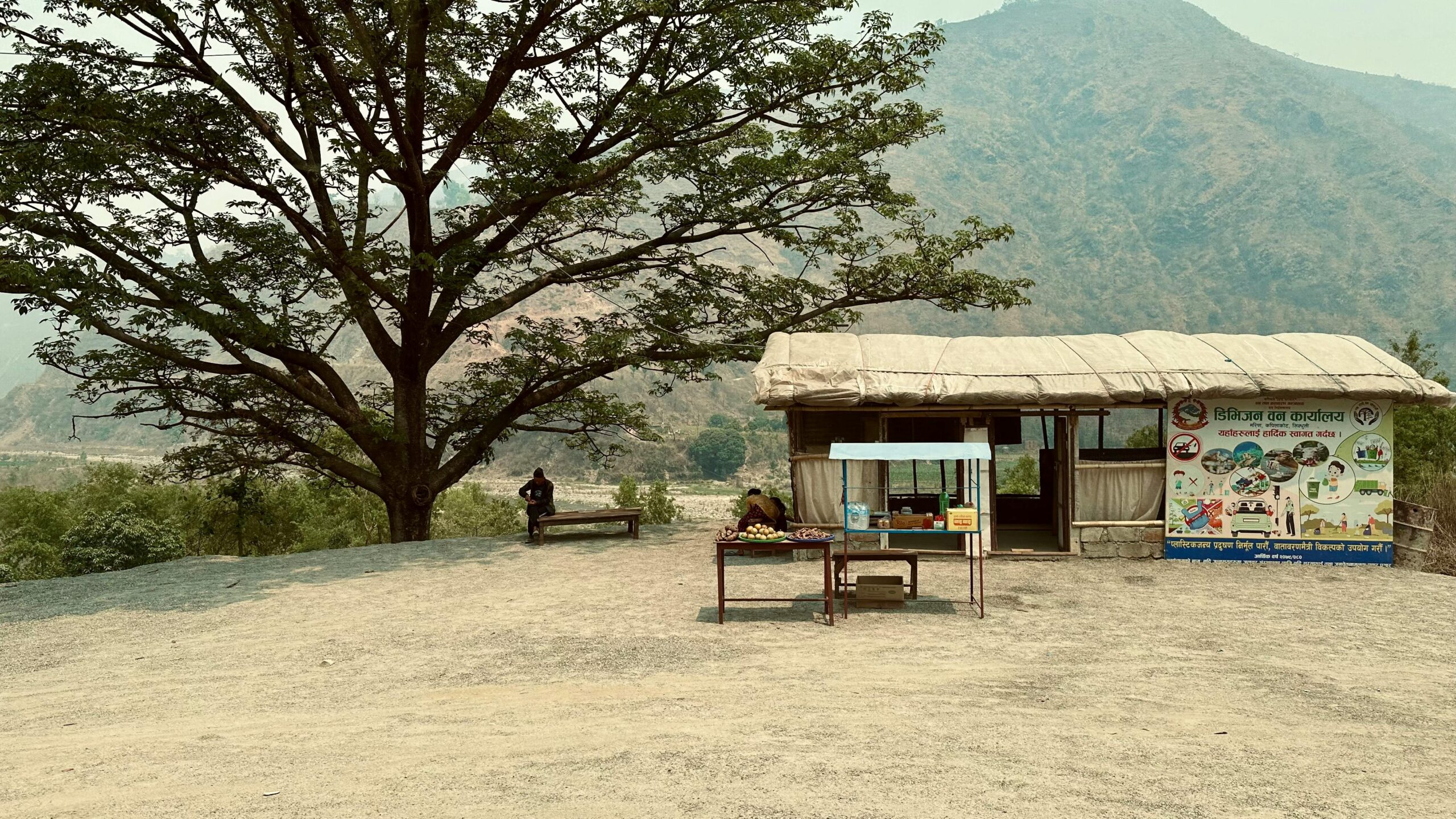 Serene mountain landscape in Nepal with a rustic bench and hut under a large tree.