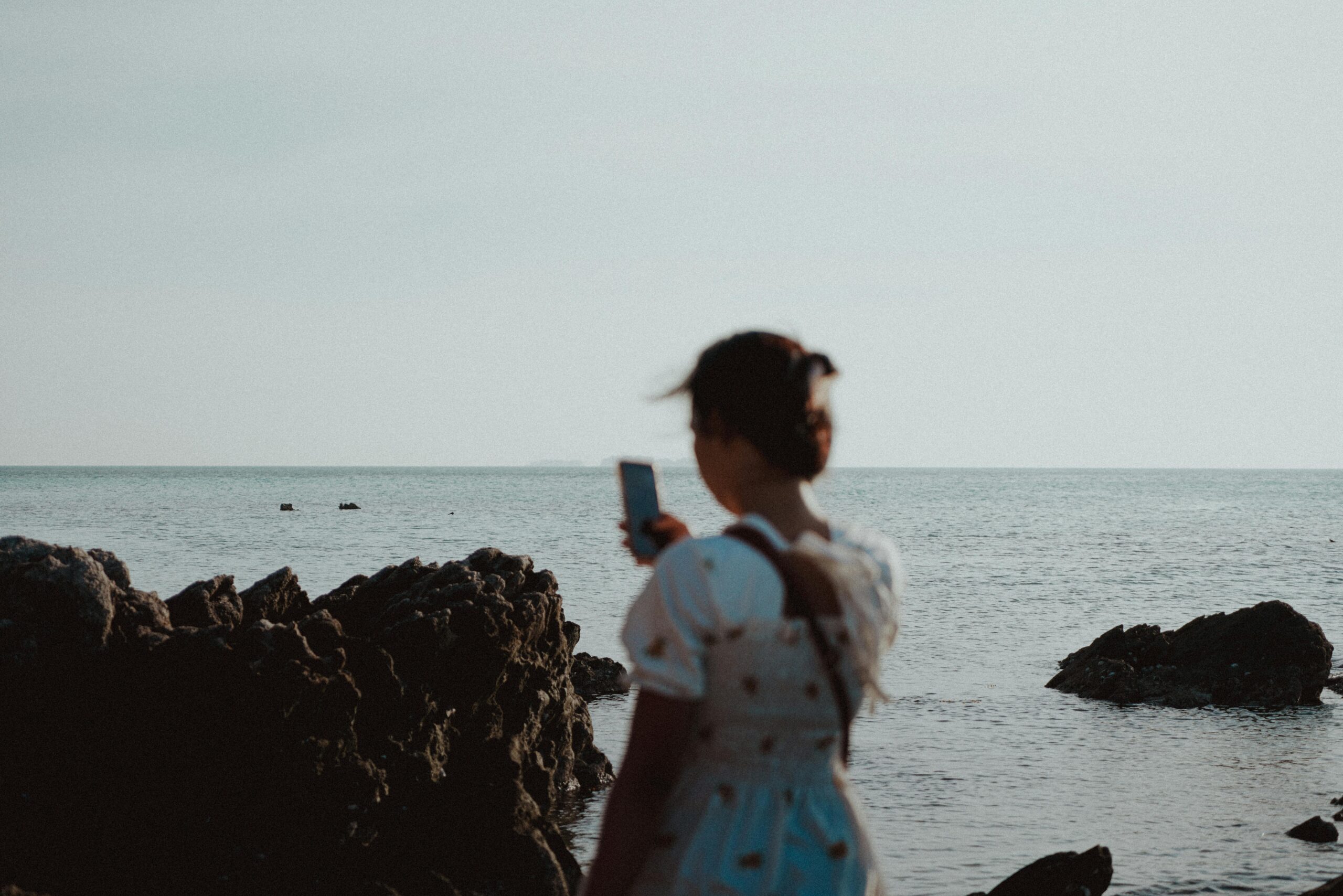 A woman stands by a rocky seaside, capturing the moment with her smartphone.