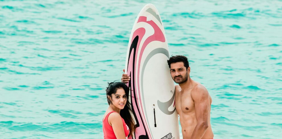 Stylish couple poses with a surfboard on a tropical Maldives beach, showcasing vibrant island fashion.