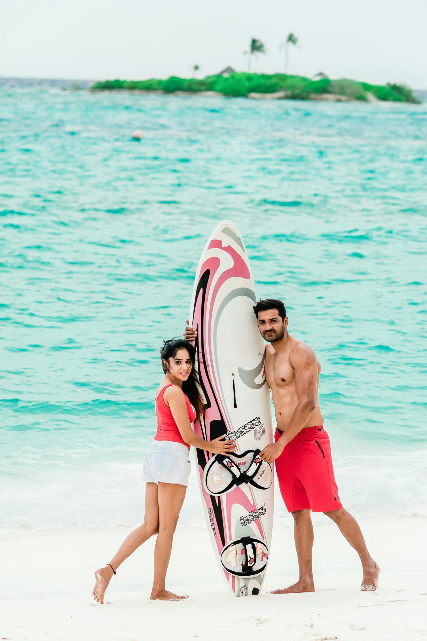 Stylish couple poses with a surfboard on a tropical Maldives beach, showcasing vibrant island fashion.