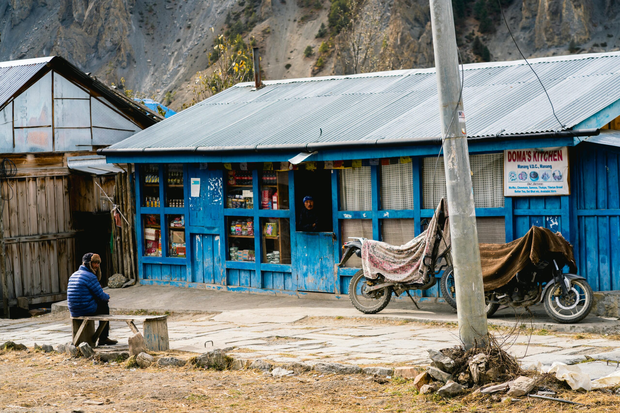 A serene scene at a rustic shop in Samagaun, Nepal, with a solitary figure outside and Himalayan landscape.