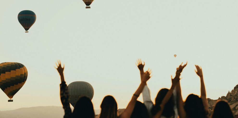 Silhouettes of women watching hot air balloons at sunset, arms raised in excitement.