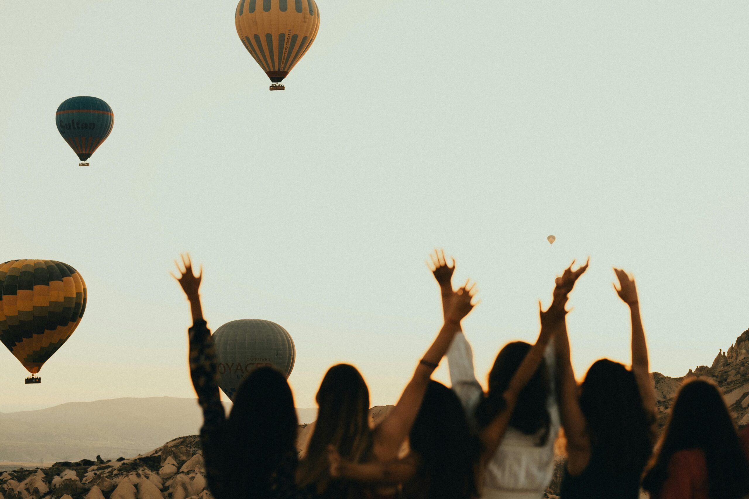 Silhouettes of women watching hot air balloons at sunset, arms raised in excitement.