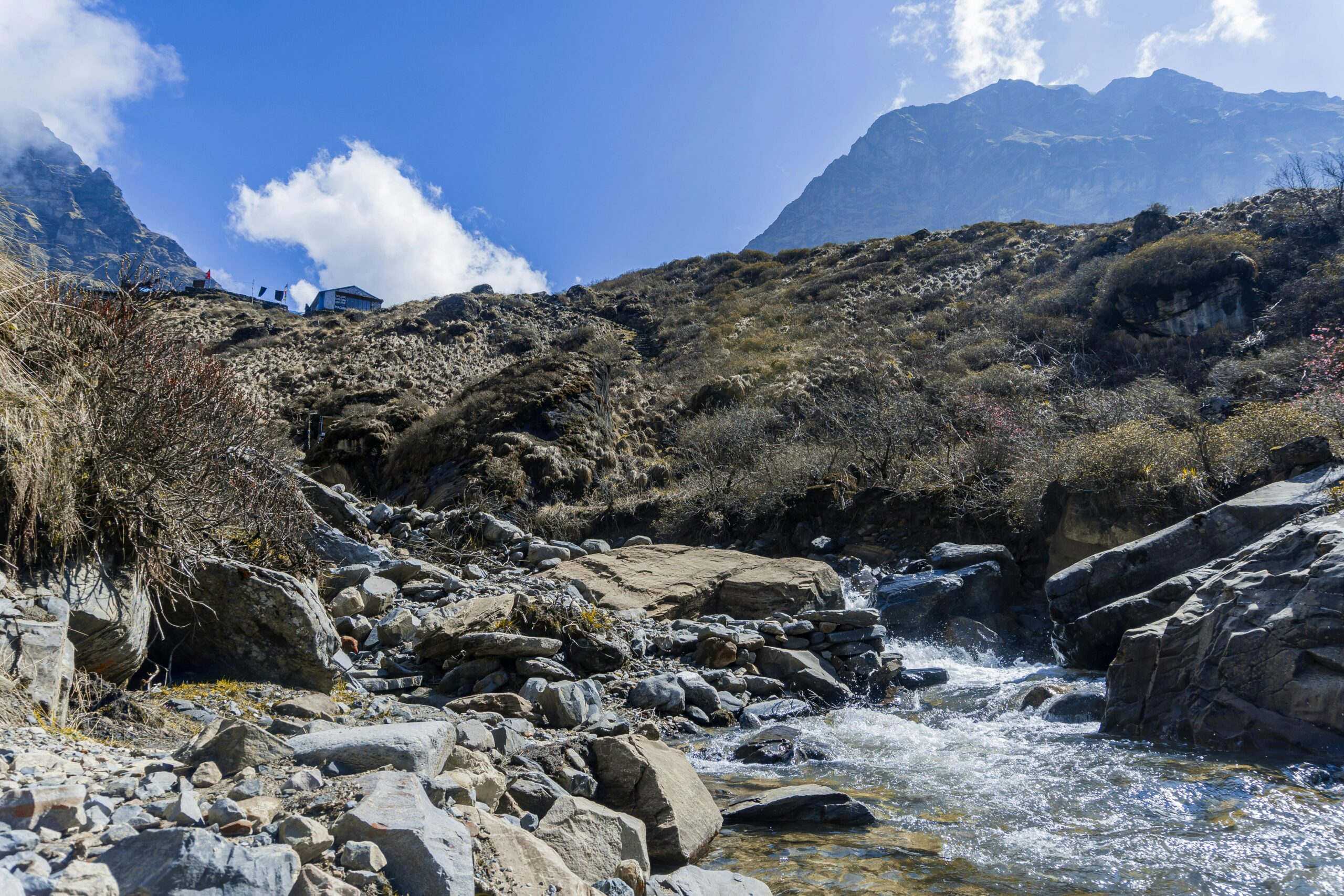 Capture of a scenic mountain stream in Ghandruk, Nepal, showcasing rugged terrain and clear blue sky.
