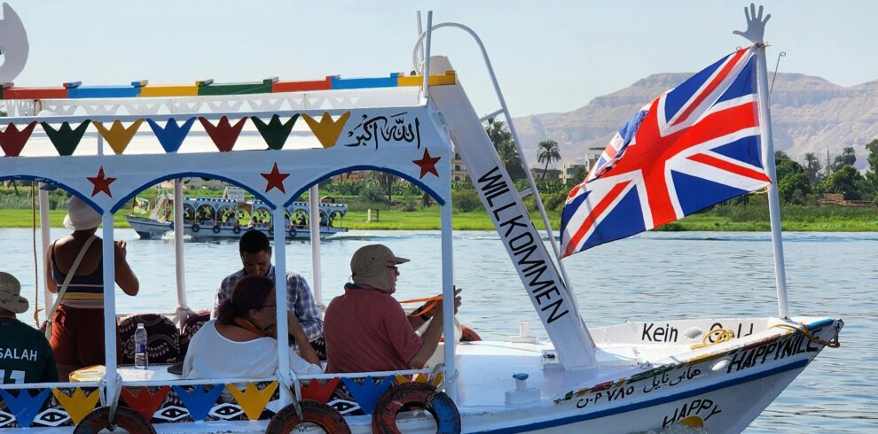 A vibrant boat on the Nile with a Union Jack flag and passengers observing the scenic view.