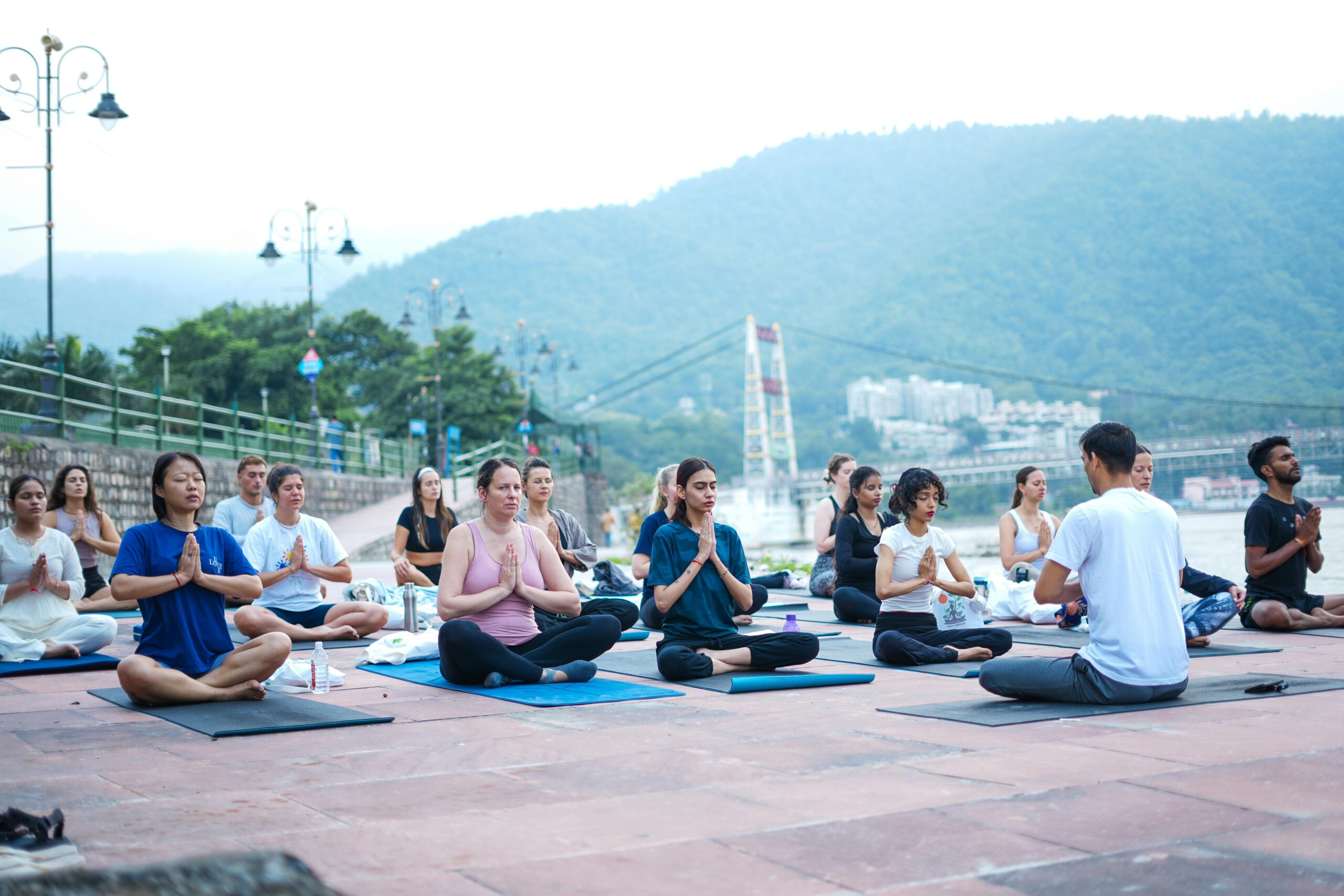 A diverse group practicing yoga by the river in Rishikesh, India, surrounded by scenic mountains.