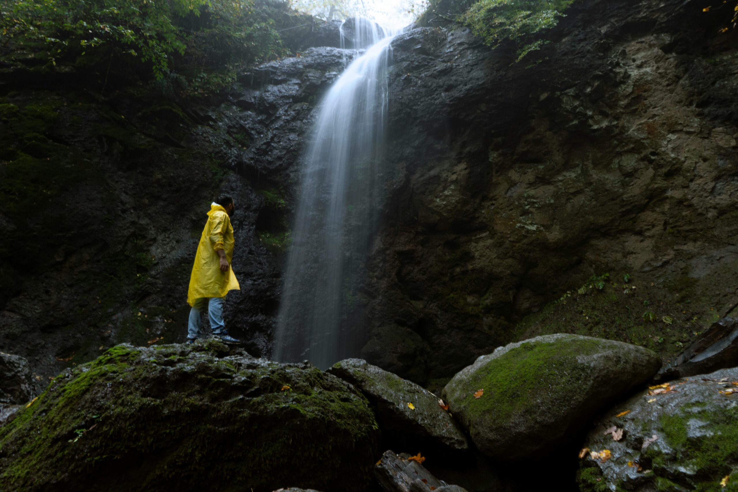 An adventurer in a yellow raincoat stands by a waterfall in a lush forest.