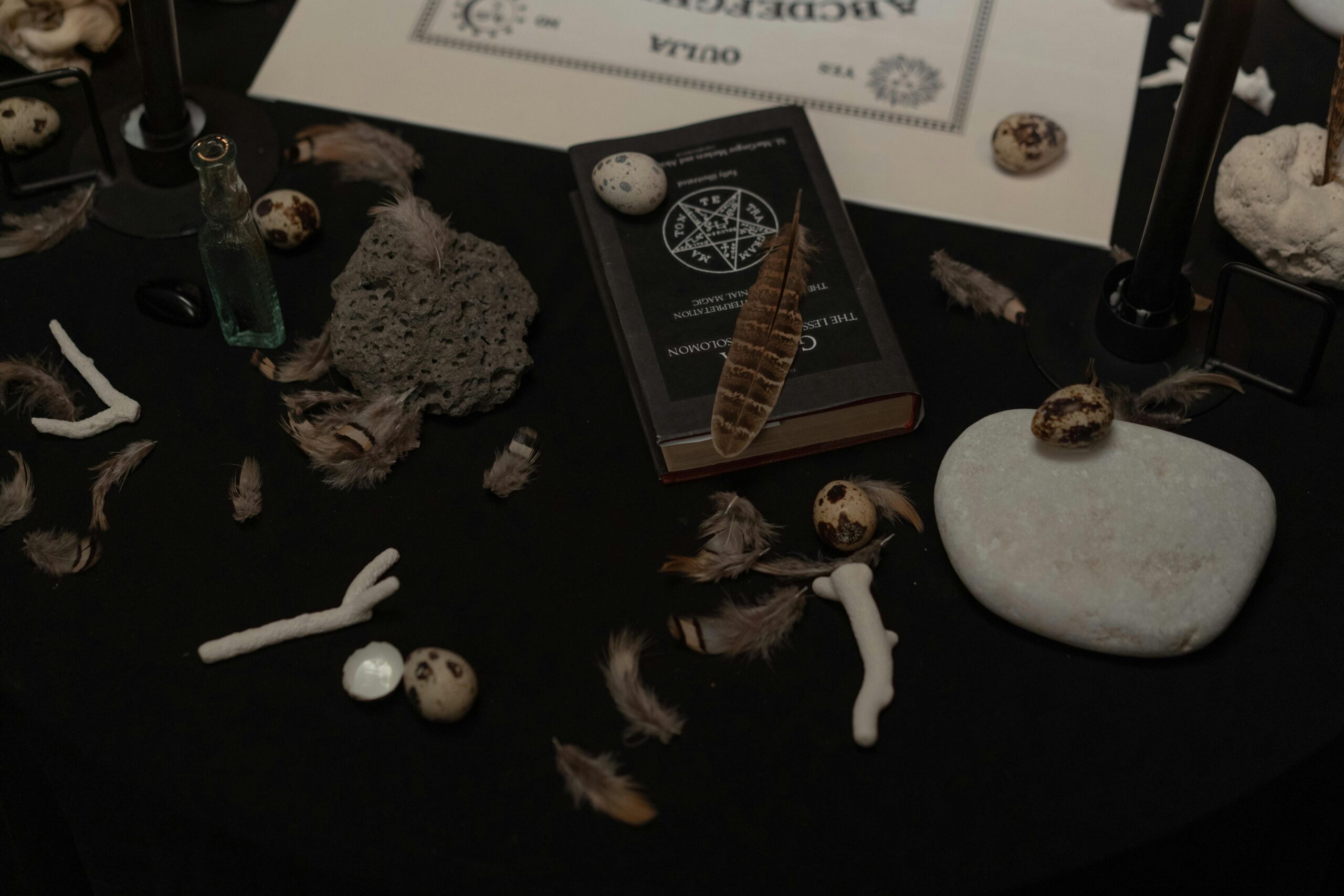 A dark, mysterious scene with a book, feathers, stones, and ritualistic items.
