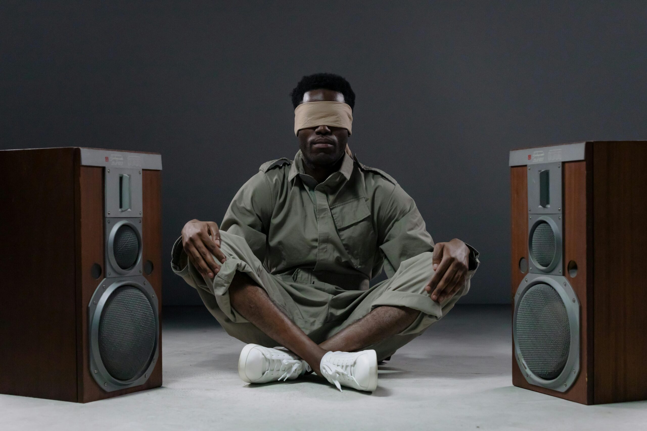 Blindfolded man in green outfit sitting between large speakers in a minimalist setting.