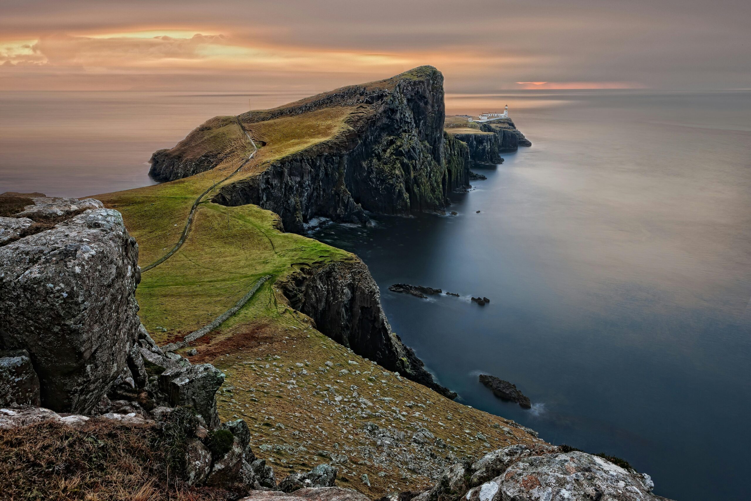 A stunning sunset view of Neist Point Lighthouse with cliffs and serene sea in Scotland.