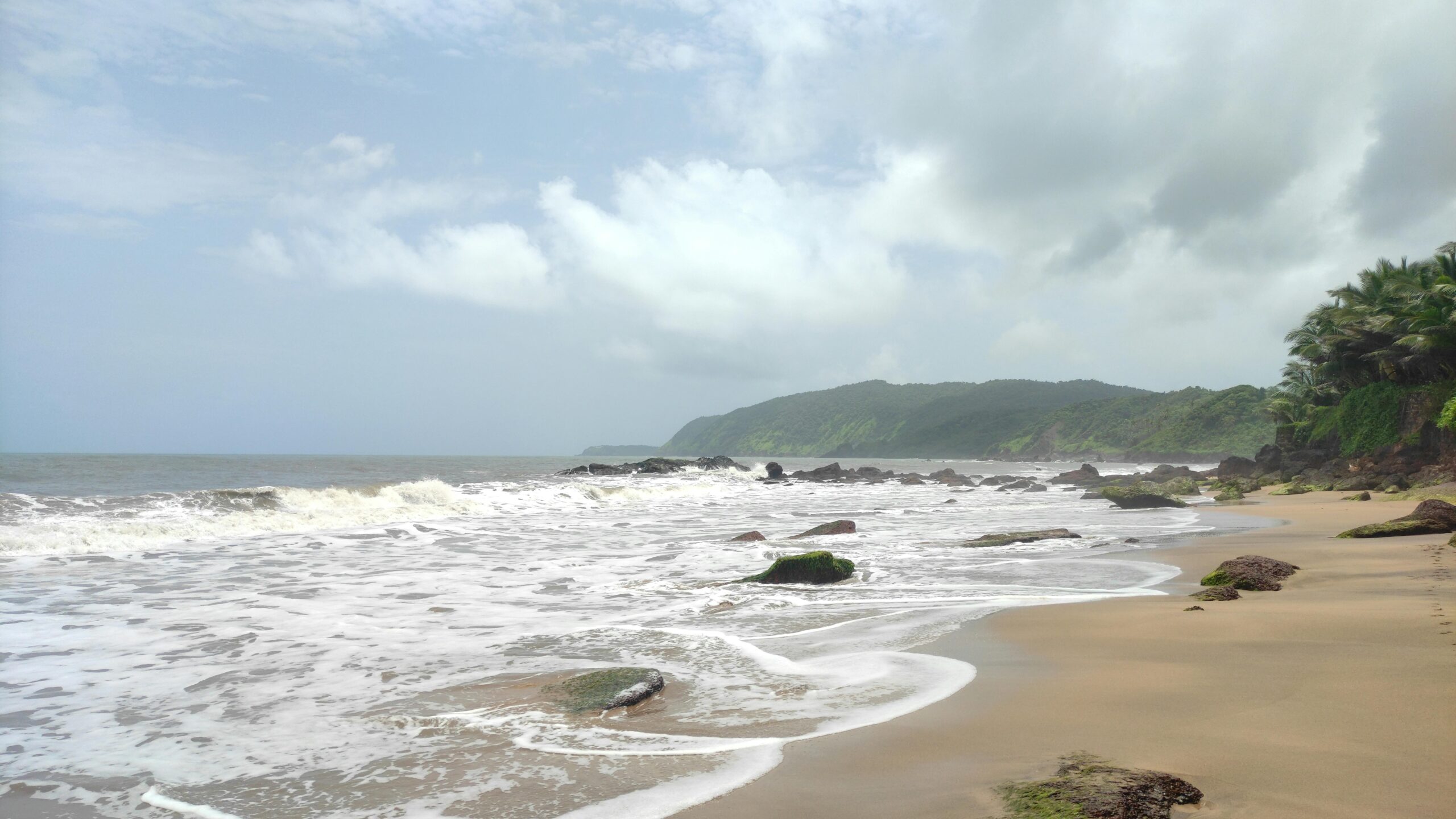 Peaceful view of Panaji beach with waves crashing on the rocks under a cloudy sky.