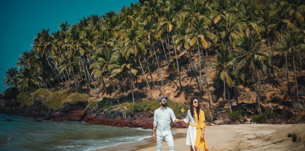 Couple enjoying a sunny beach walk in Goa with lush palm trees and clear blue waters.